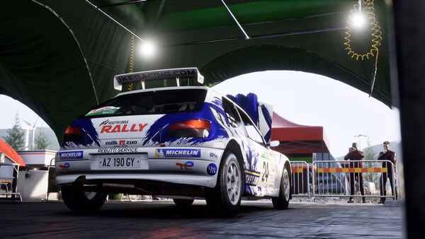 Screenshot from Assetto Corsa Rally: A rally car is parked under a green canopy in a service area.